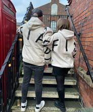 Load image into Gallery viewer, A guy and girl on some stairs in amazing oversized hoodies! The hoodies are off-white and have a bold back DEFIANT print in black that spans both arms and the top of the back.  There is also embroidery on the back that reads &#39;Proudly Refusing To Obey Authority&#39; with 2018 in Roman numerals underneath that.  The hoodies are designed to be quite oversized to provide you with that cosy feel.