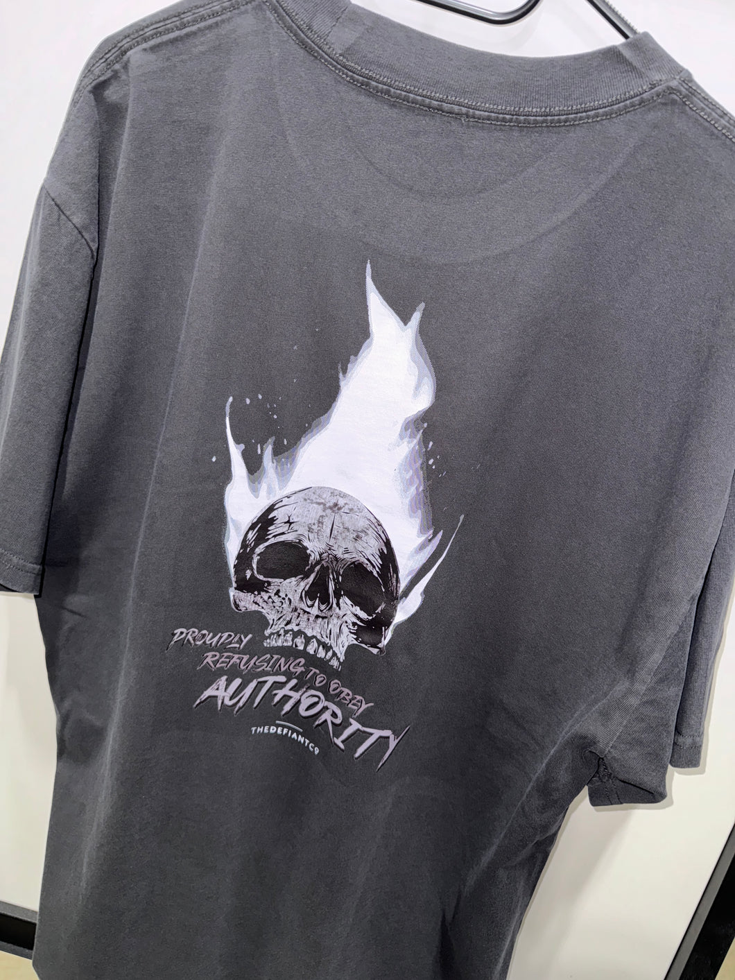 A photo showing the back of a unisex t-shirt.  The shirt has a big bold print on the back of a skull in flames. Underneath the skull is the defiant tag line 'Proudly Refusing To Obey Authority' with the The Defiant Co logo under that. The t-shirt is faded coal in colour.