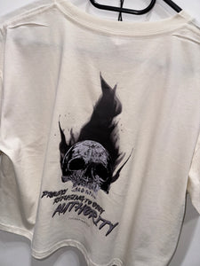 A photo showing the back of a female fit cropped t-shirt.  The shirt has a big bold print on the back of a skull in flames. Underneath the slull is the defiant tag line 'Proudly Refusing To Obey Authority' with the The Defiant Co logo under that. The shirt is ecru in colour.