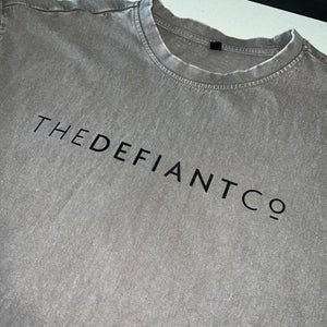 A photo showing an oversized The Defiant Co T-Shirt.  The shirt has the famous ‘The Defiant Co’ logo across the front of the chest.  The shirt has a round neck and is oversized.  This particular version has a washed finish and is the colour asphalt with black print.