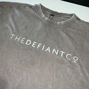 A photo showing an oversized The Defiant Co T-Shirt.  The shirt has the famous ‘The Defiant Co’ logo across the front of the chest.  The shirt has a round neck and is oversized.  This particular version has a washed finish and is the colour asphalt with white print.