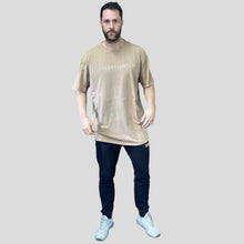 Load image into Gallery viewer, A photo showing a guy wearing an oversized The Defiant Co T-Shirt.  The shirt has the famous ‘The Defiant Co’ logo across the front of the chest.  The shirt has a round neck and is oversized.  This particular version has a washed finish and is the colour Washed Beige.