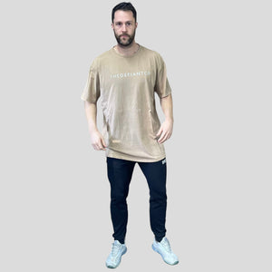 A photo showing a guy wearing an oversized The Defiant Co T-Shirt.  The shirt has the famous ‘The Defiant Co’ logo across the front of the chest.  The shirt has a round neck and is oversized.  This particular version has a washed finish and is the colour Washed Beige.