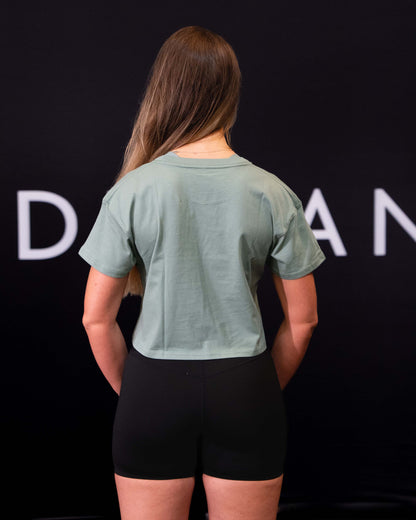 A girl against a black The Defiant Co backdrop wearing the TDCo cropped t-shirt in sage green with the black DFNT. Booty shorts. 