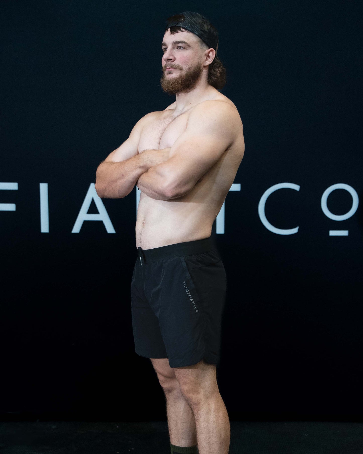 A man against a black The Defiant Co backdrop wearing the performance shorts with their draw string and elasticated black waist band.  The Defiant Co is subtly embossed down the right leg. The colour is black.