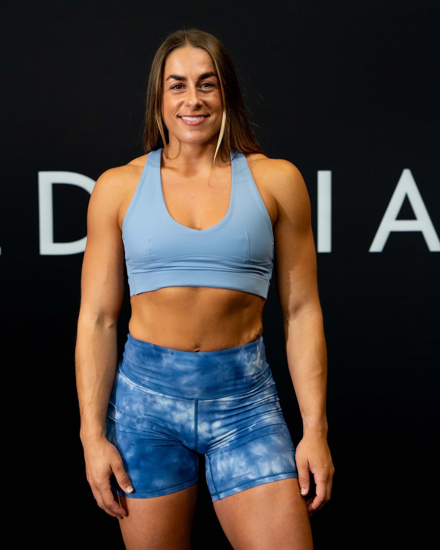 A girl against a black The Defiant Co backdrop wearing the Infinity Sports bra with it's crossed back and supportive front.  The Defiant Co is criss-crossed over both straps across the back. The colour is serene blue.
