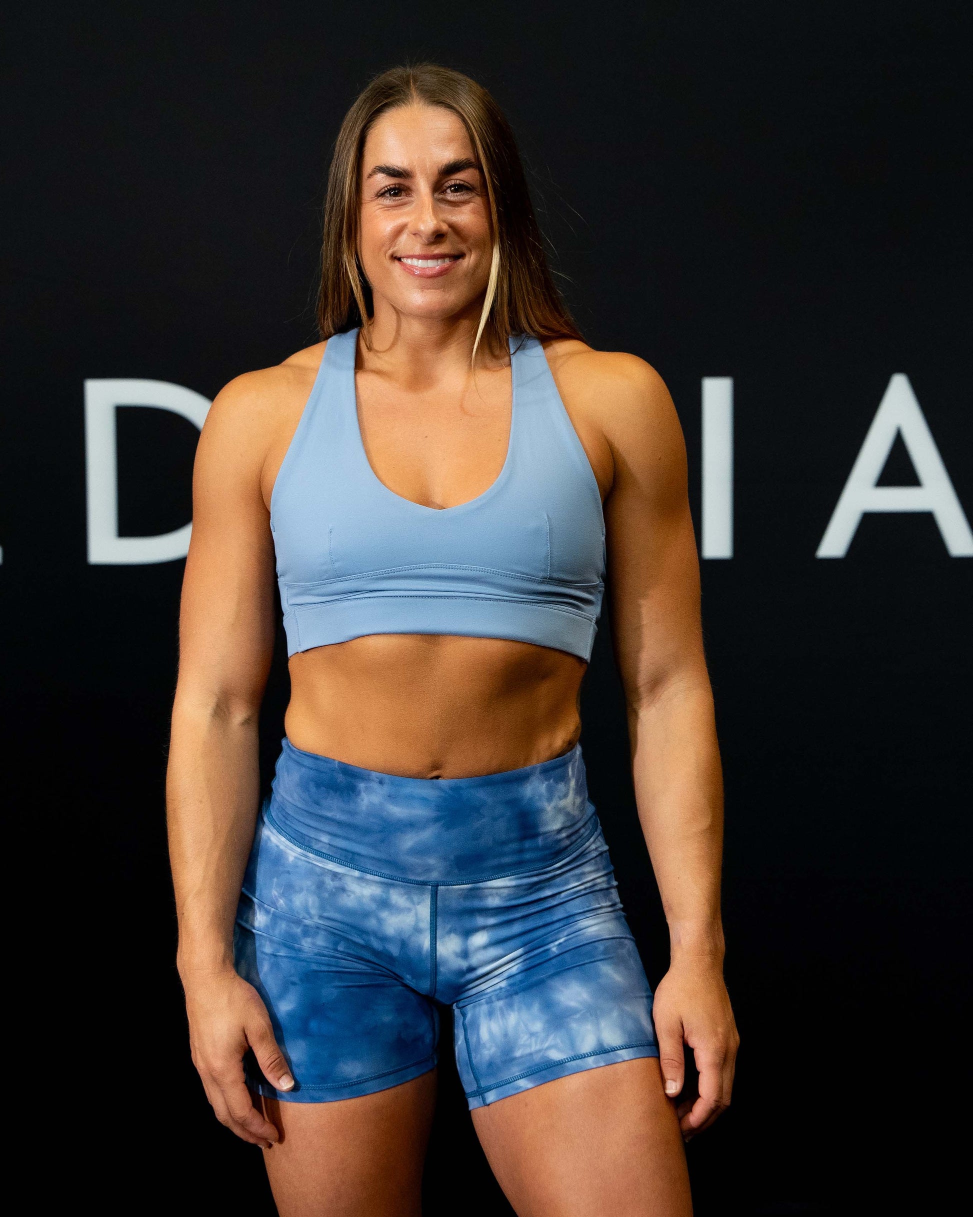 A girl against a black The Defiant Co backdrop wearing the Infinity Sports bra with it's crossed back and supportive front.  The Defiant Co is criss-crossed over both straps across the back. The colour is serene blue.