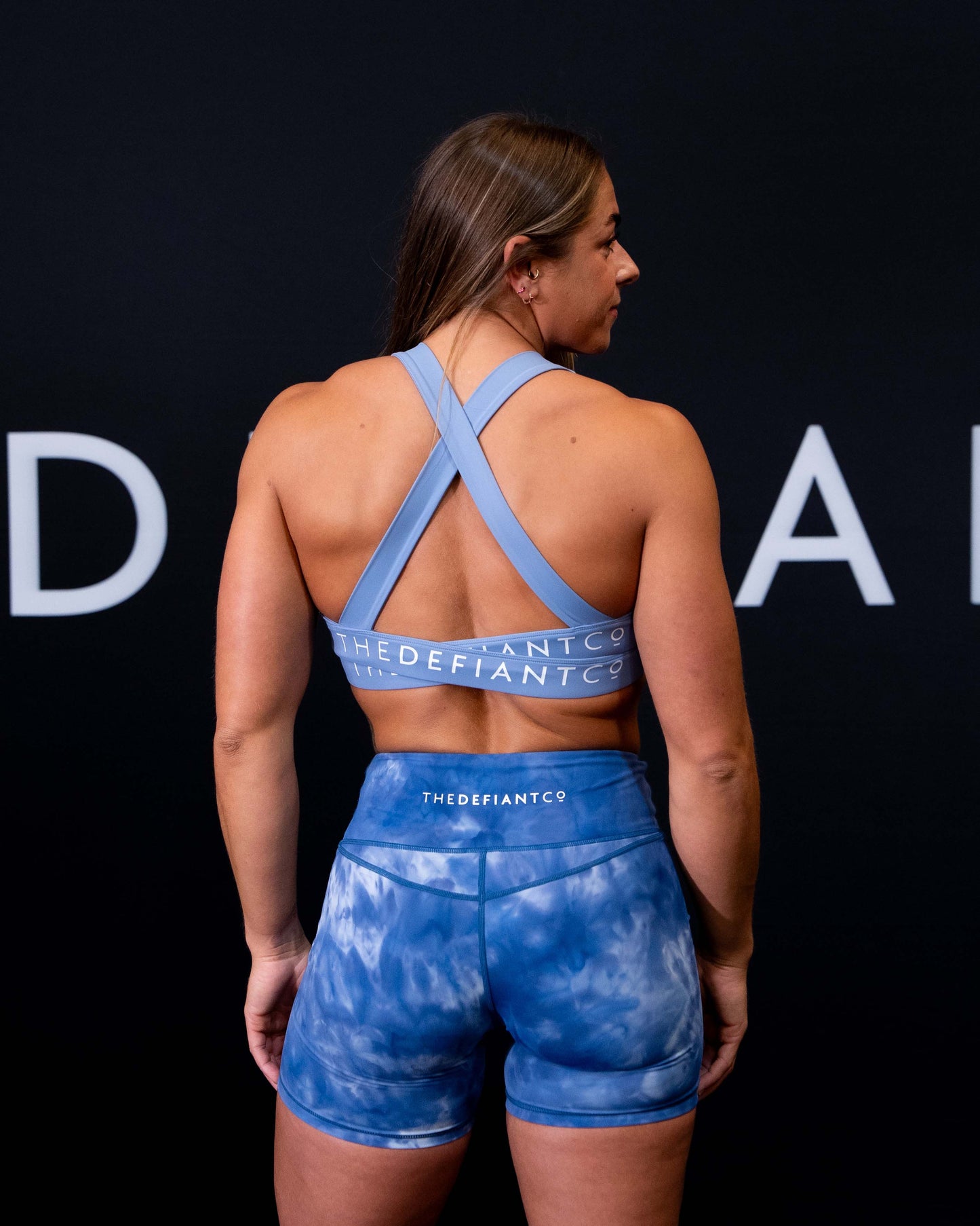 A girl against a black The Defiant Co backdrop wearing the Infinity Sports bra with it's crossed back and supportive front.  The Defiant Co is criss-crossed over both straps across the back. The colour is serene blue.
