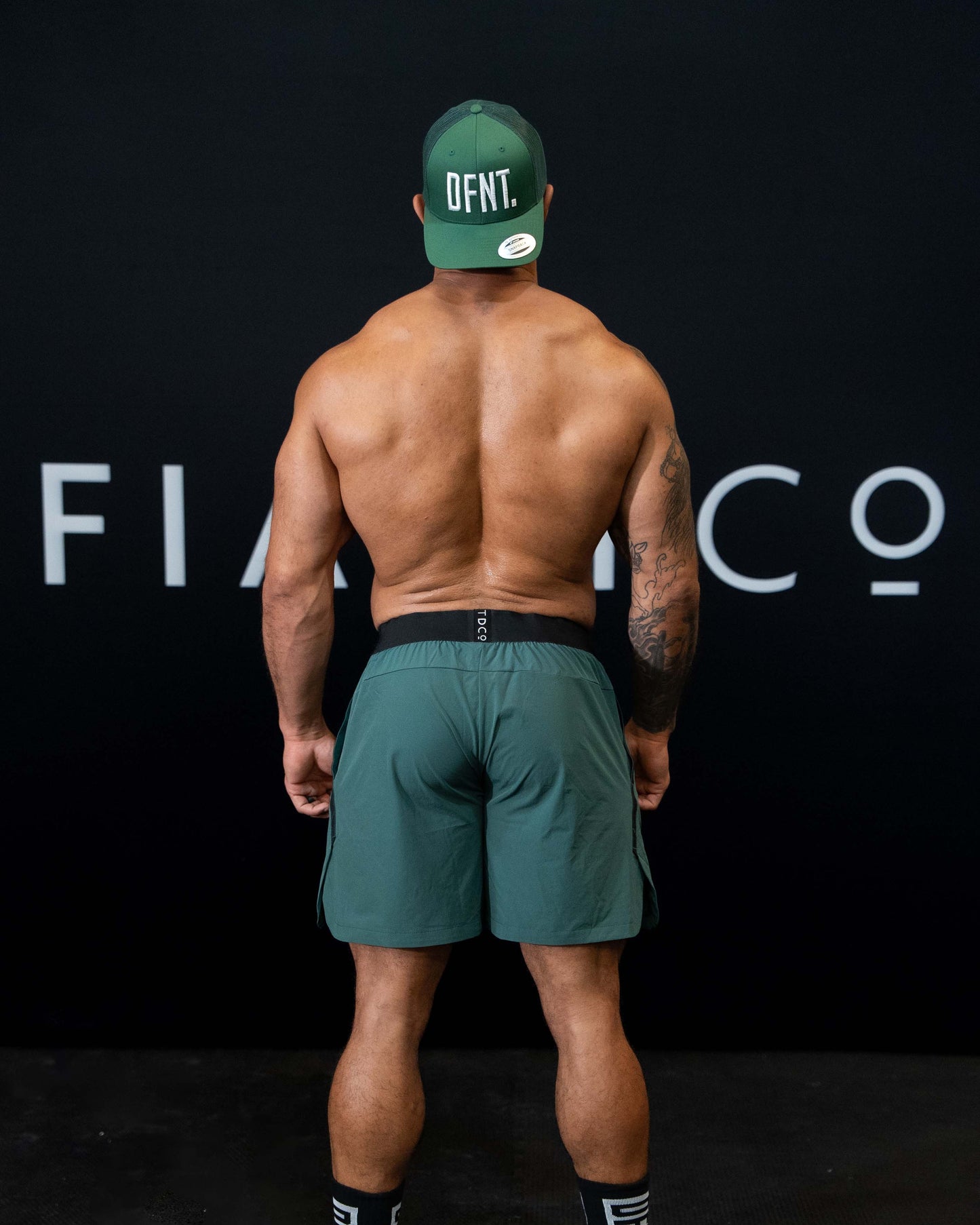 A man against a black The Defiant Co backdrop wearing the performance shorts with their draw string and elasticated black waist band.  The Defiant Co is subtly embossed down the right leg. The colour is racing green.