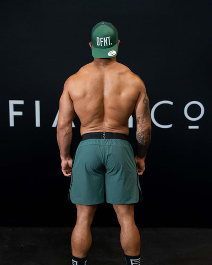 A man against a black The Defiant Co backdrop wearing the performance shorts with their draw string and elasticated black waist band.  The Defiant Co is subtly embossed down the right leg. The colour is racing green.