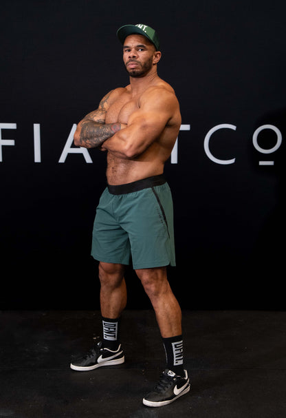 A man against a black The Defiant Co backdrop wearing the performance shorts with their draw string and elasticated black waist band.  The Defiant Co is subtly embossed down the right leg. The colour is racing green.