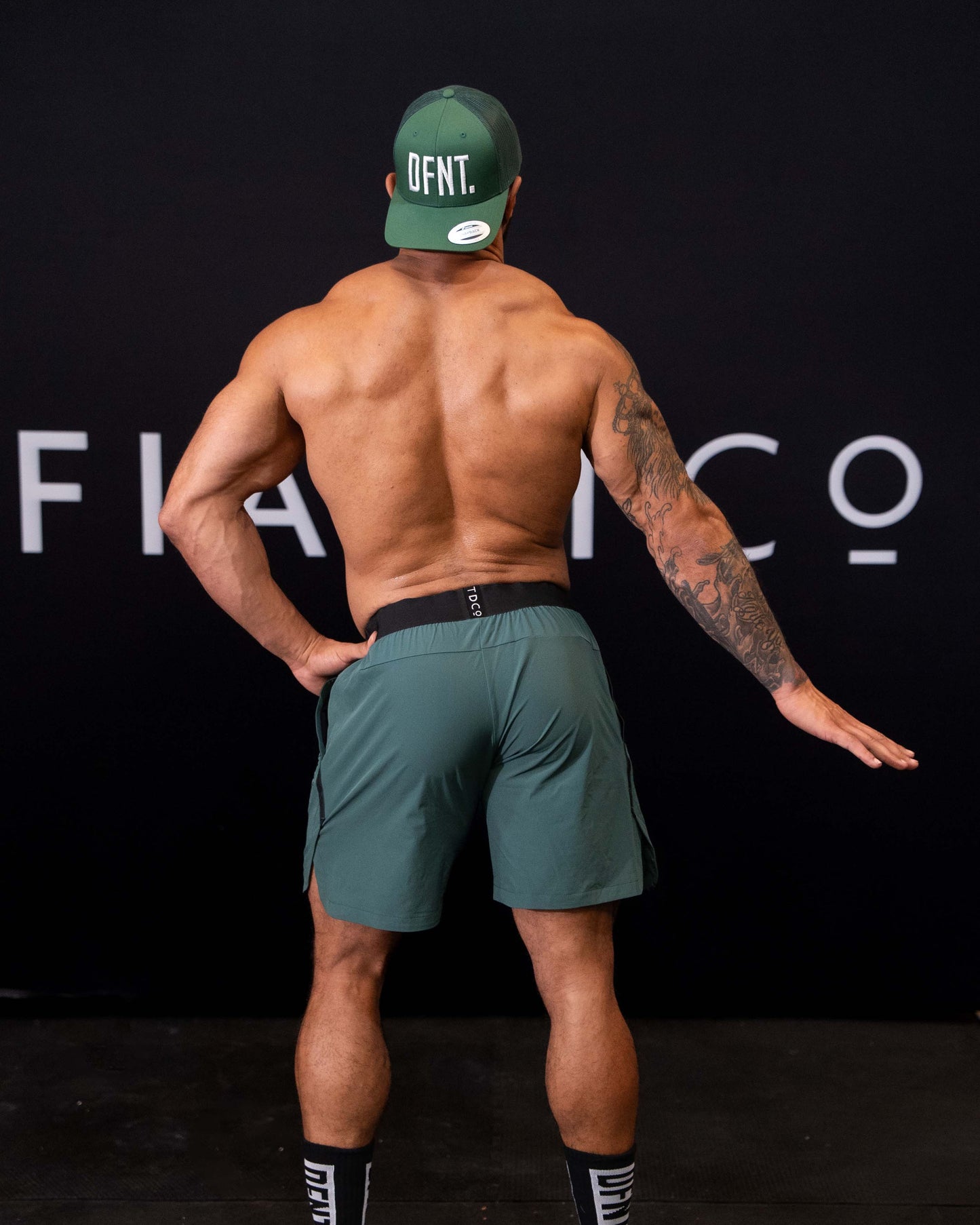 A man against a black The Defiant Co backdrop wearing the performance shorts with their draw string and elasticated black waist band.  The Defiant Co is subtly embossed down the right leg. The colour is racing green.