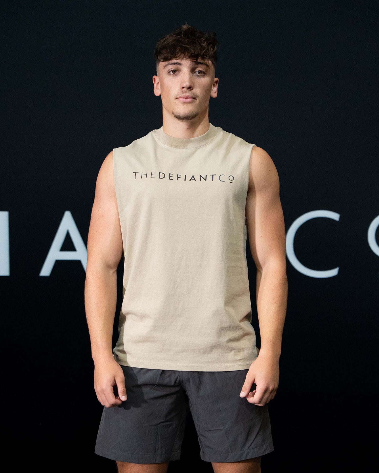 A guy against a black The Defiant Co backdrop wearing the TDCo sand sleeveless tee, charcoal performance shorts.