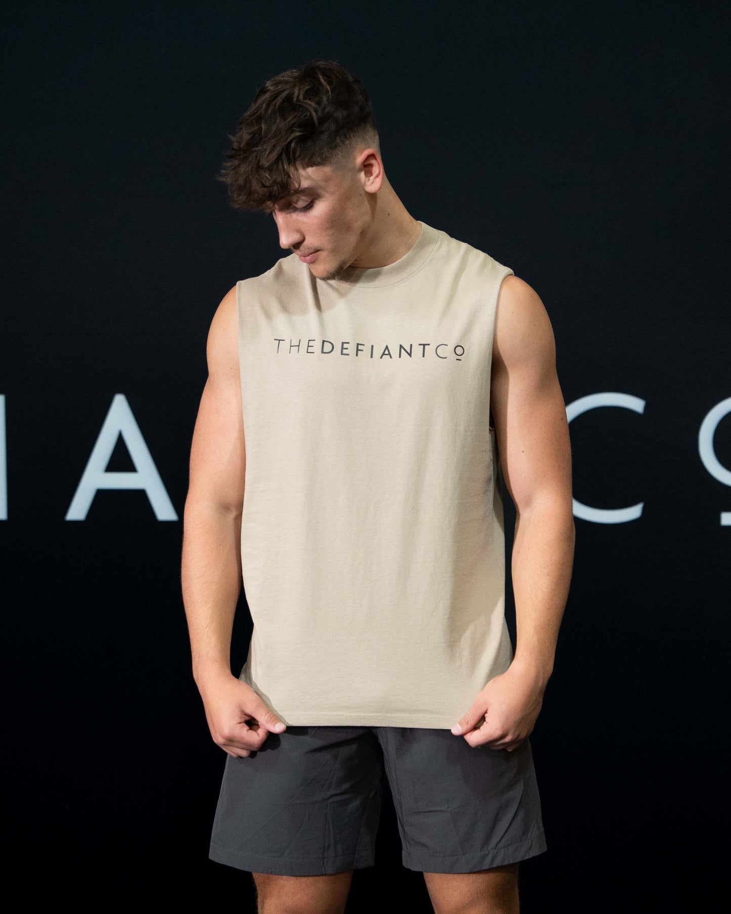 A guy against a black The Defiant Co backdrop wearing the TDCo sand sleeveless tee, charcoal performance shorts.