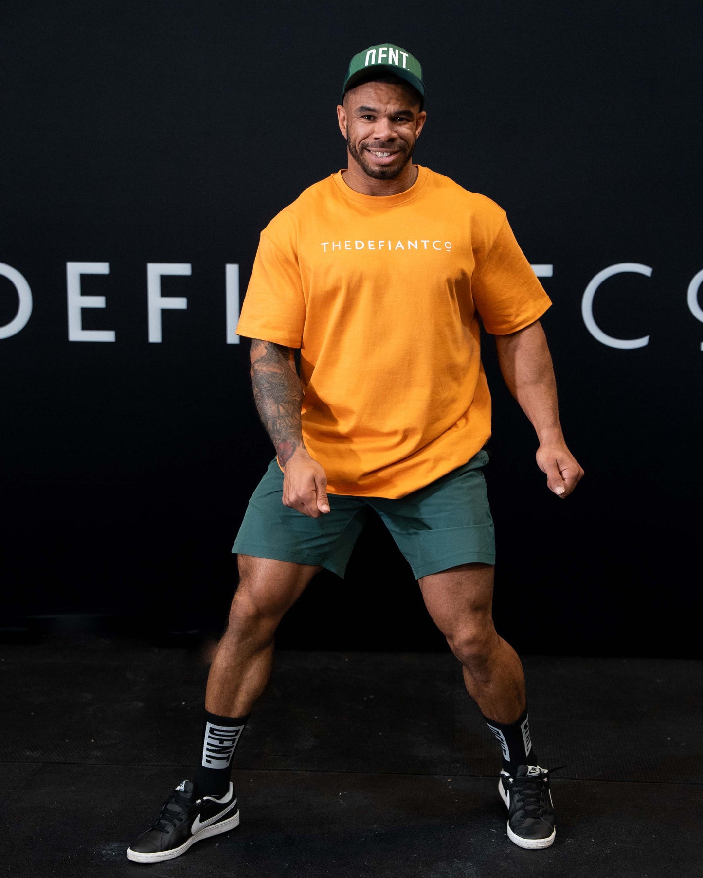 A man against a black The Defiant Co backdrop wearing the DFNT. Bottle Green Trucker hat, Bottle green TDCo performance shorts and Orange TDCo t-shirt.