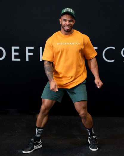 A man against a black The Defiant Co backdrop wearing the DFNT. Bottle Green Trucker hat, Bottle green TDCo performance shorts and Orange TDCo t-shirt.