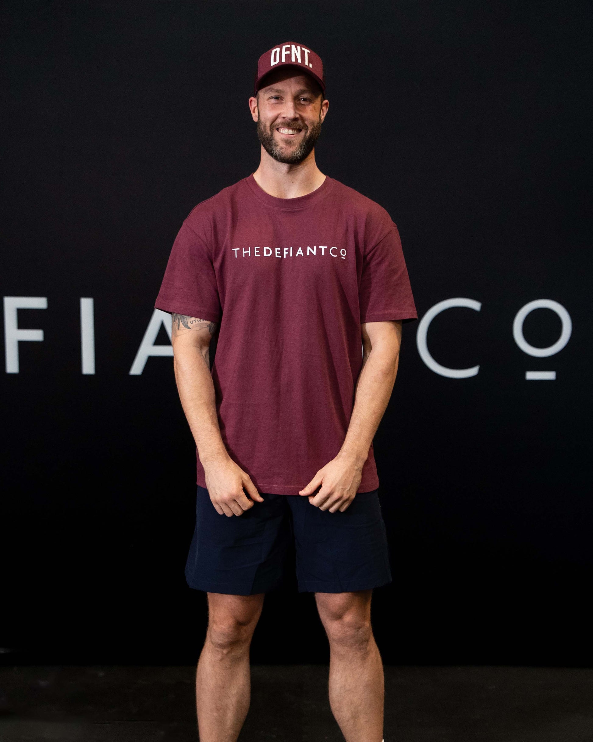 A man against a black The Defiant Co backdrop wearing the DFNT. Burgundy Trucker hat, Navy TDCo performance shorts and Burgundy TDCo tee.