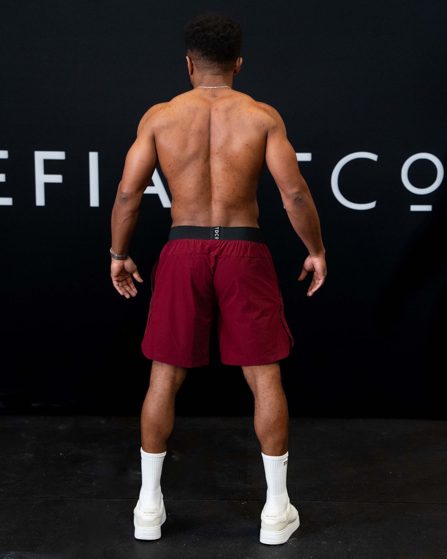 A man against a black The Defiant Co backdrop wearing the performance shorts with their draw string and elasticated black waist band.  The Defiant Co is subtly embossed down the right leg. The colour is burgundy