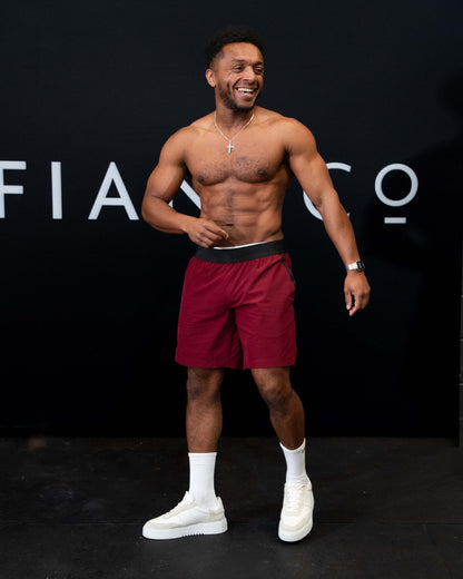 A man against a black The Defiant Co backdrop wearing the performance shorts with their draw string and elasticated black waist band.  The Defiant Co is subtly embossed down the right leg. The colour is burgundy.