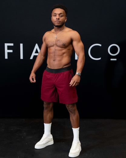 A man against a black The Defiant Co backdrop wearing the performance shorts with their draw string and elasticated black waist band.  The Defiant Co is subtly embossed down the right leg. The colour is burgundy