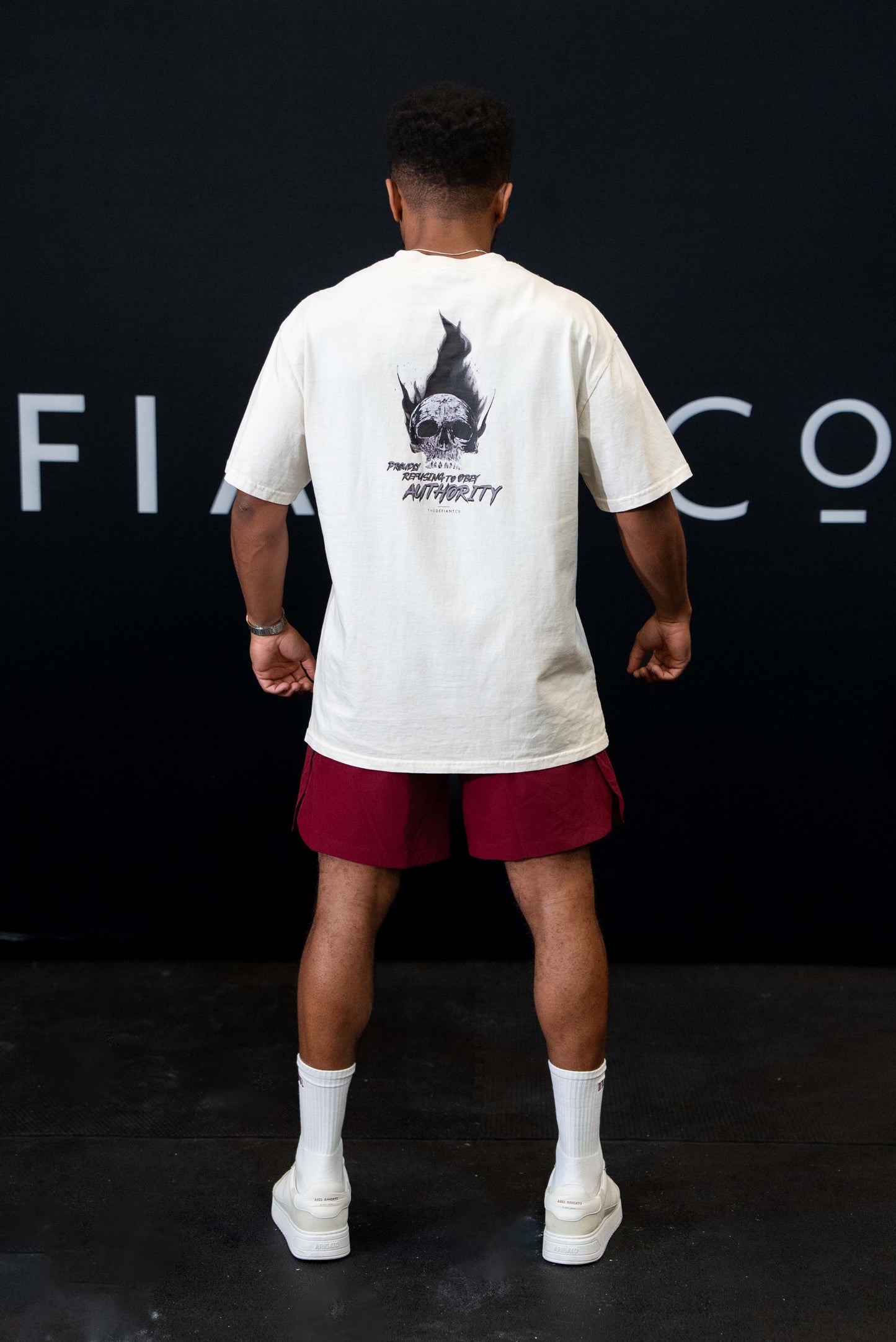 A guy against a black The Defiant Co backdrop wearing the faded ecru proudly refusing to obey authority oversized tee and burgundy performance shorts. 