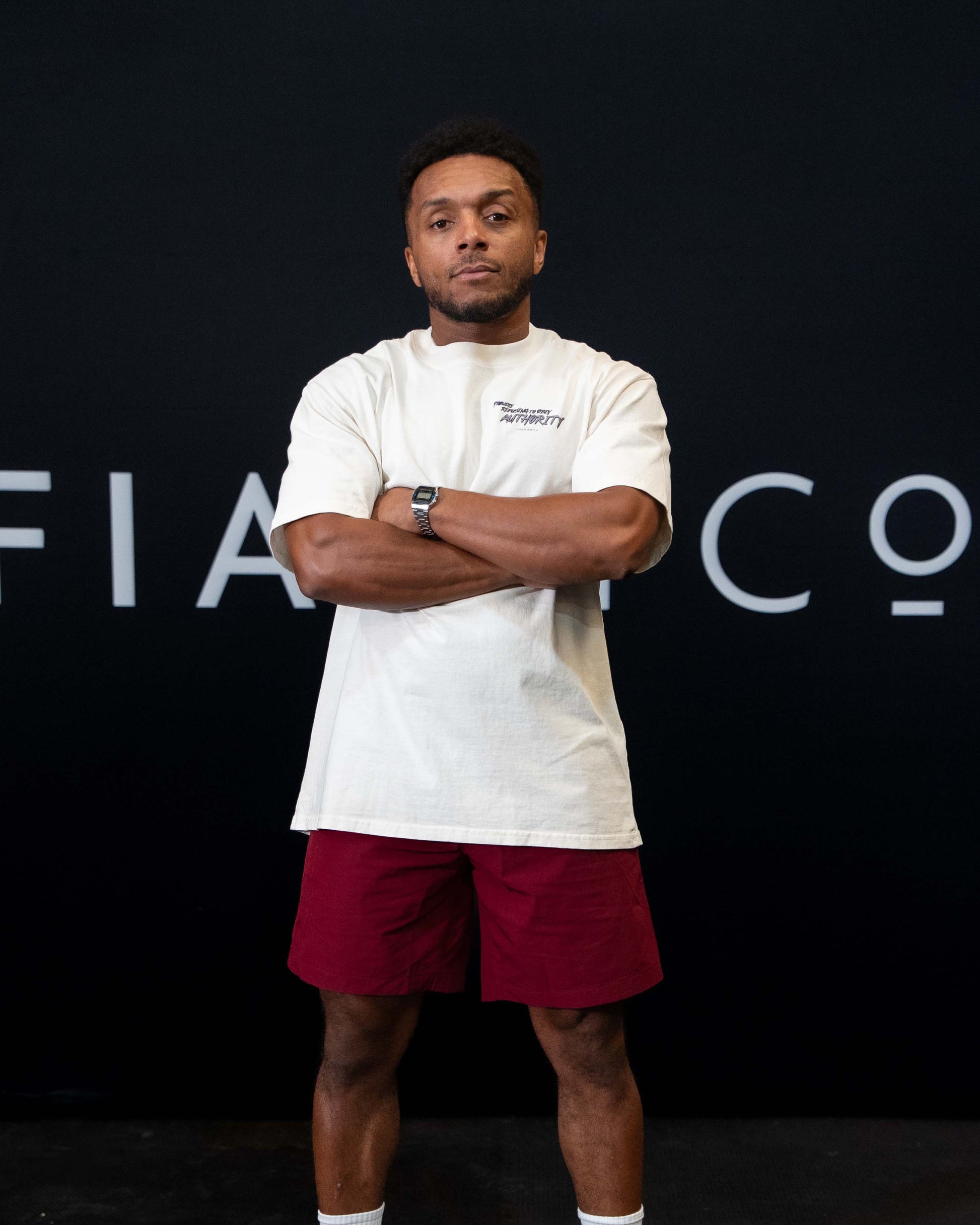 A guy against a black The Defiant Co backdrop wearing the faded ecru proudly refusing to obey authority oversized tee and burgundy performance shorts. 