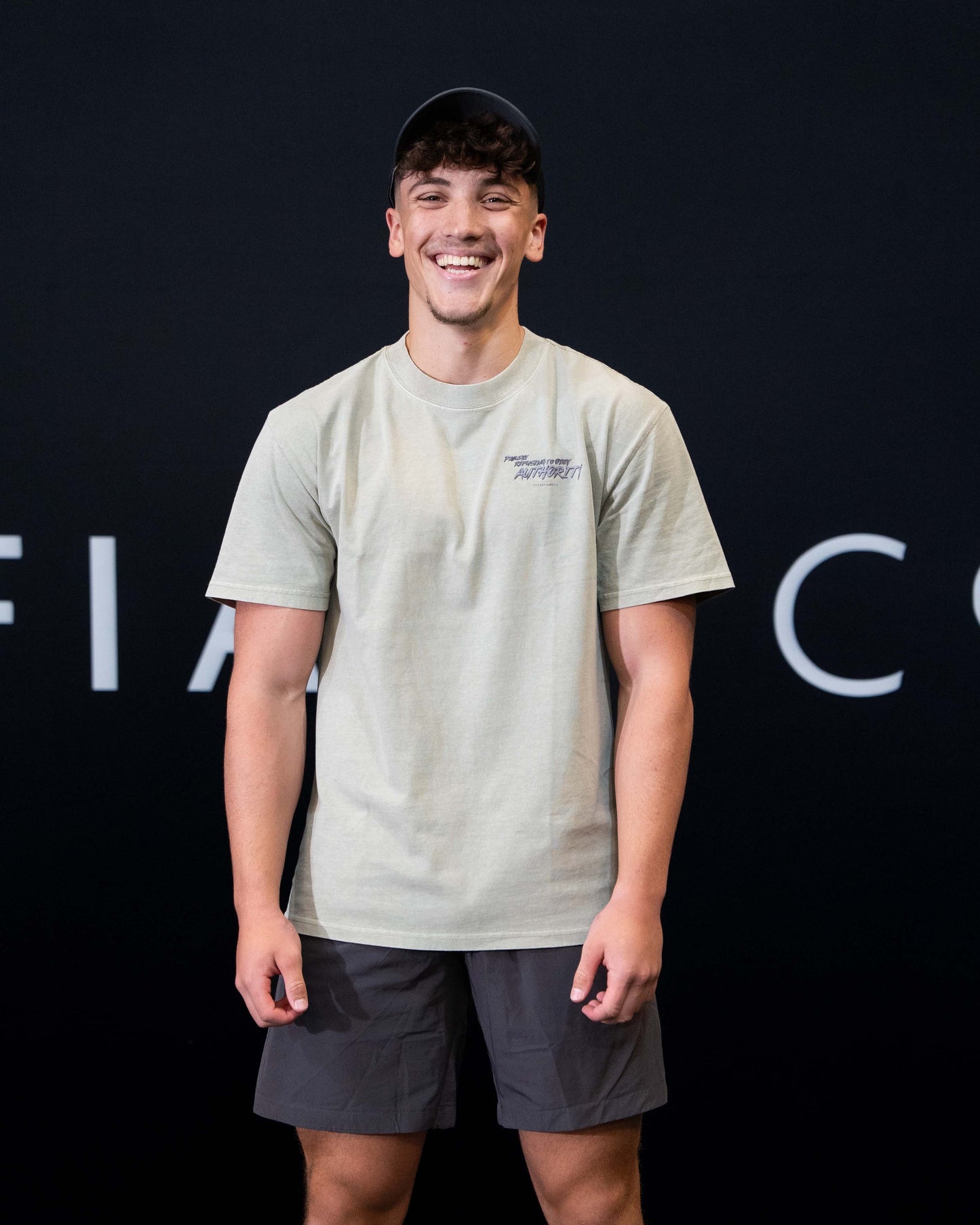 A guy against a black The Defiant Co backdrop wearing the faded khaki proudly refusing to obey authority oversized tee and charcoal performance shorts. 