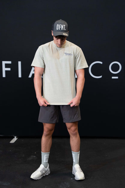 A man against a black The Defiant Co backdrop wearing the DFNT. Charcoal Trucker hat, charcoal TDCo performance shorts and faded khaki proudly refusing to obey authority t-shirt.