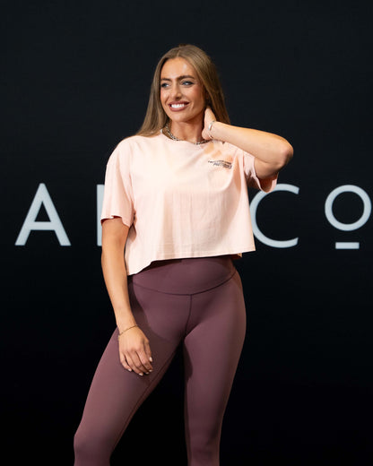 A girl against a black The Defiant Co backdrop wearing the proudly refusing to obey authority cropped t-shirt in blush with the burgundy TDCo performance leggings.