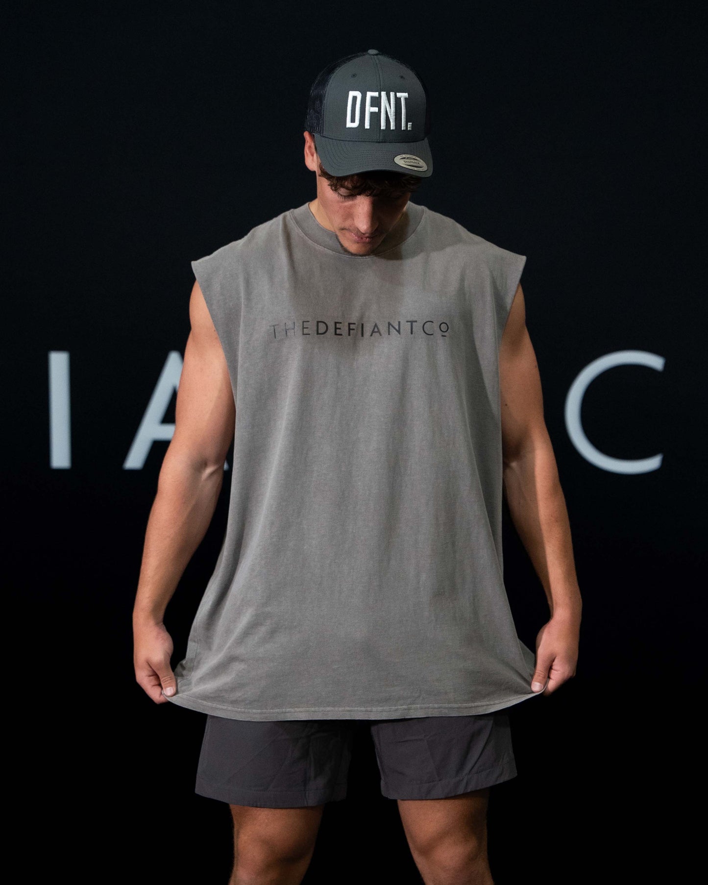 A guy against a black The Defiant Co backdrop wearing the TDCo cwashed grey sleeveless tee, charcoal performance shorts and a charcoal trucker DFNT. hat.