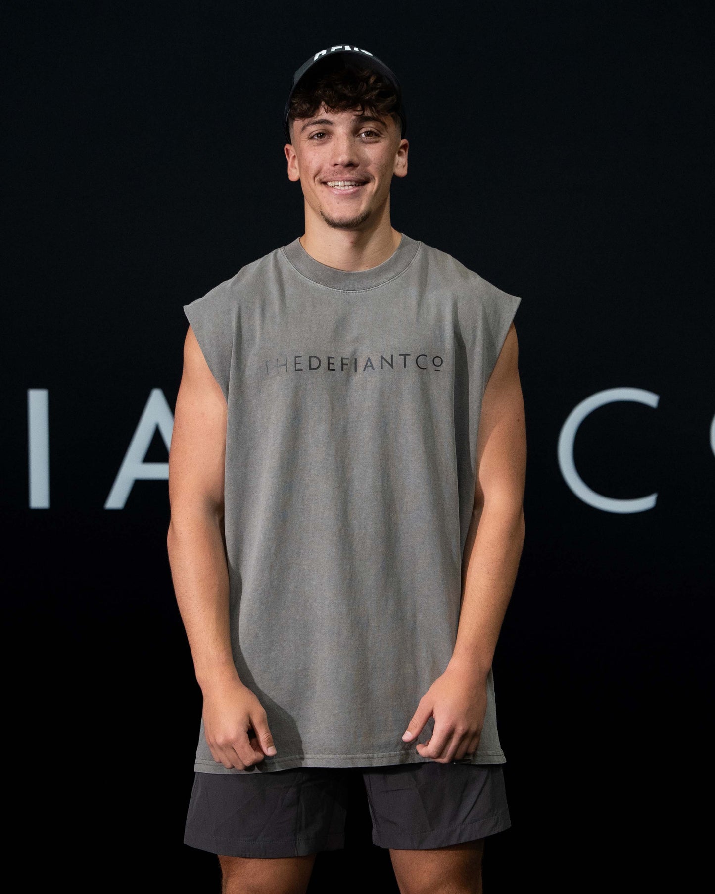 A guy against a black The Defiant Co backdrop wearing the TDCo cwashed grey sleeveless tee, charcoal performance shorts and a charcoal trucker DFNT. hat.