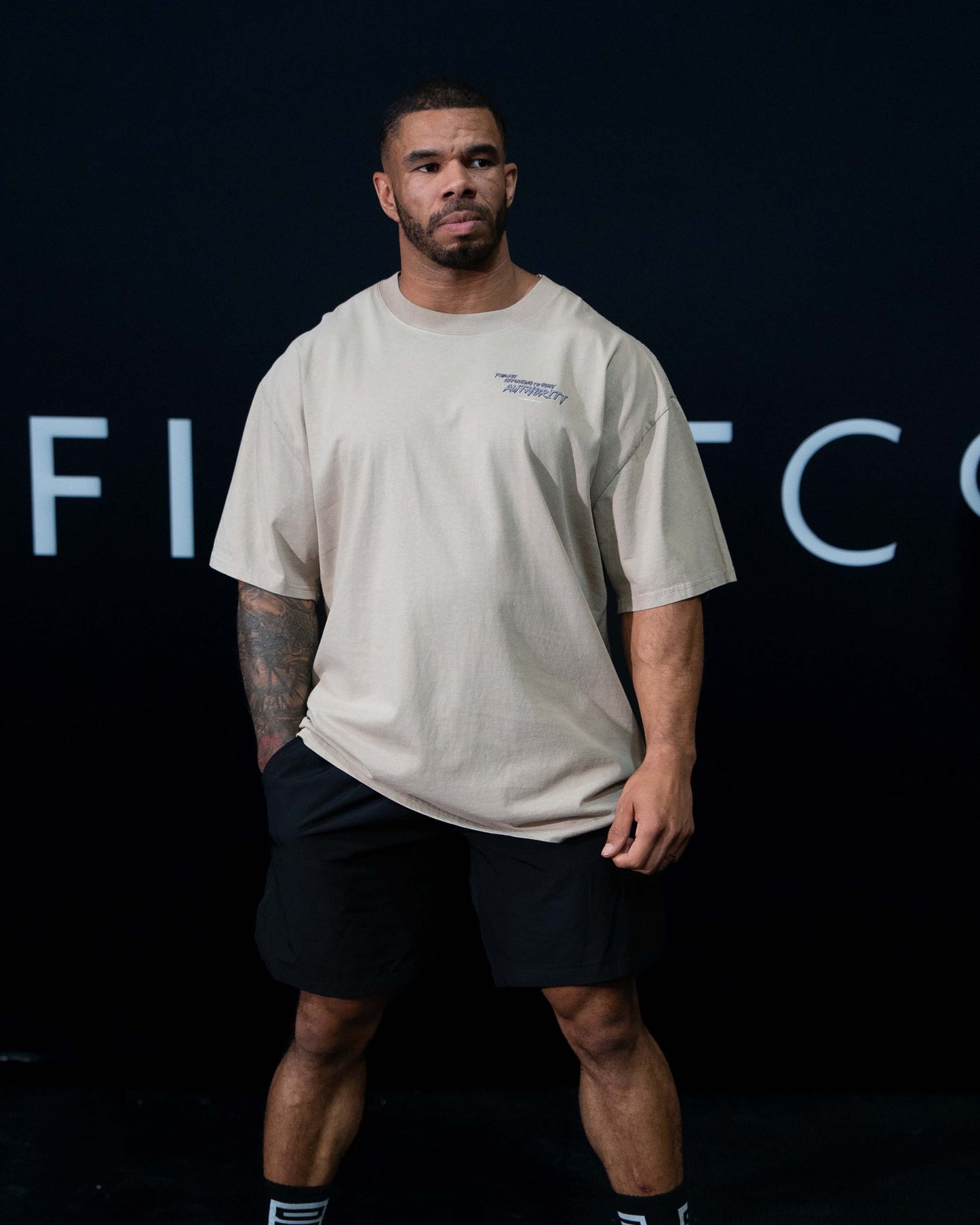 A guy against a black The Defiant Co backdrop wearing the faded khaki proudly refusing to obey authority oversized tee and black performance shorts. 