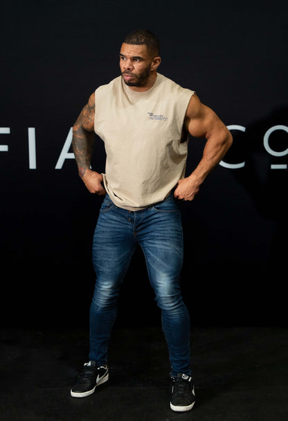 A guy against a black The Defiant Co backdrop wearing the faded khaki proudly refusing to obey authority sleeveless tee and jeans and trainers. 