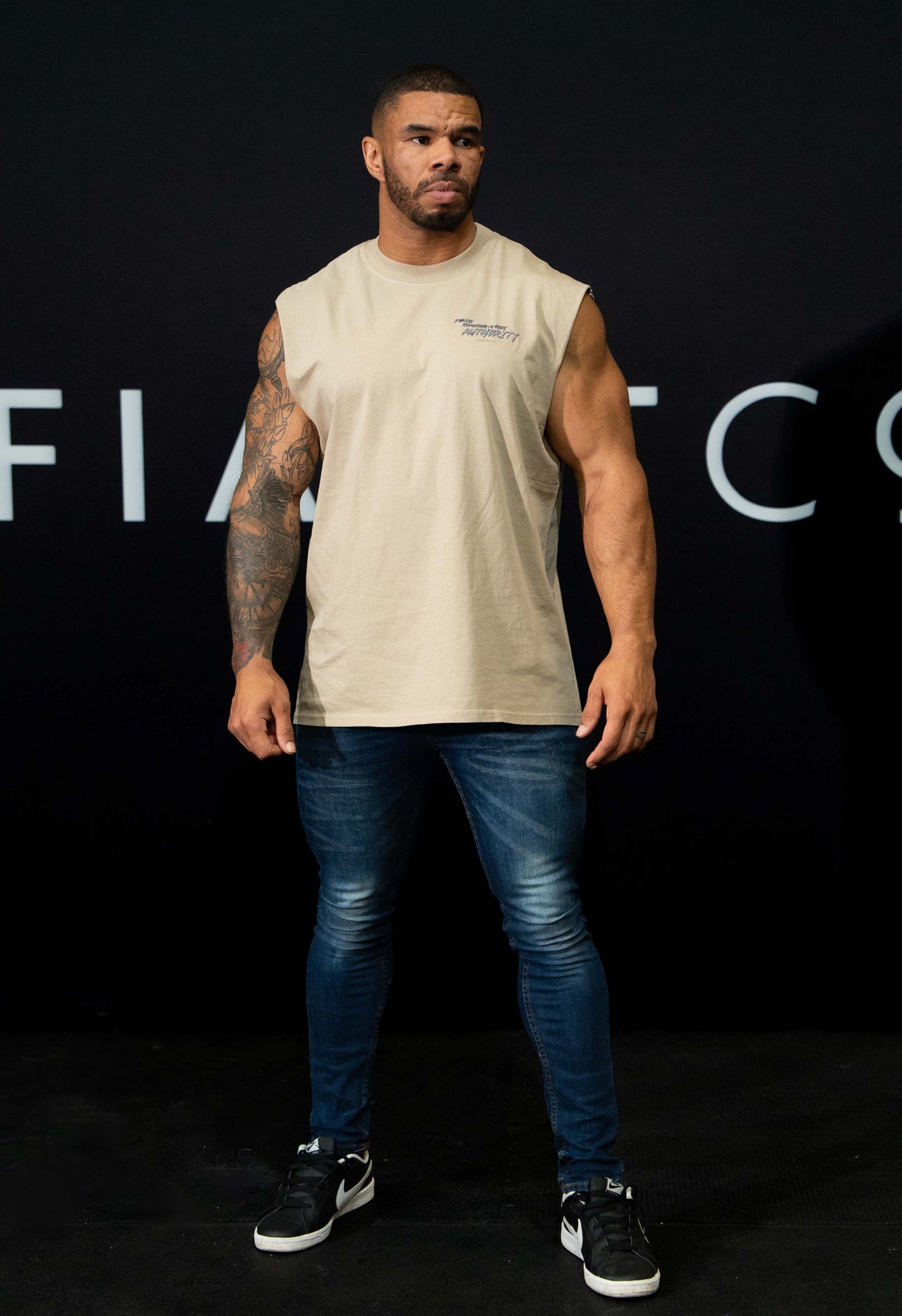 A guy against a black The Defiant Co backdrop wearing the faded khaki proudly refusing to obey authority sleeveless tee and jeans and trainers. 