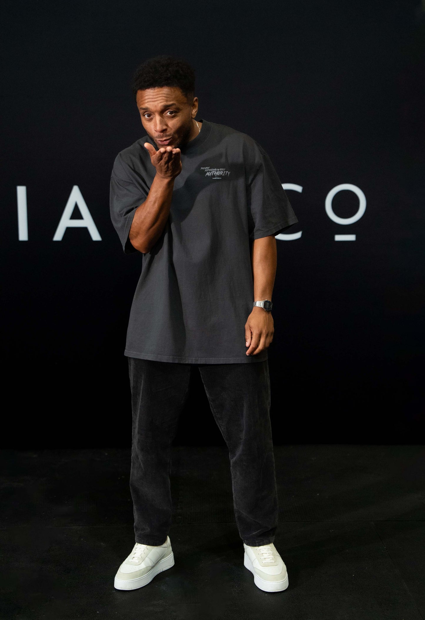 A guy against a black The Defiant Co backdrop wearing the faded black proudly refusing to obey authority oversized tee and black corduroy trousers. 