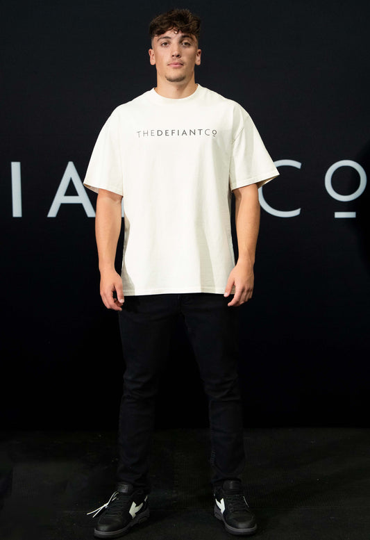 A guy against a black The Defiant Co backdrop wearing the oversized t-shirt.  The tee has the famous 'The Defiant Co' logo boldly displayed across the front. The colour is ecru.