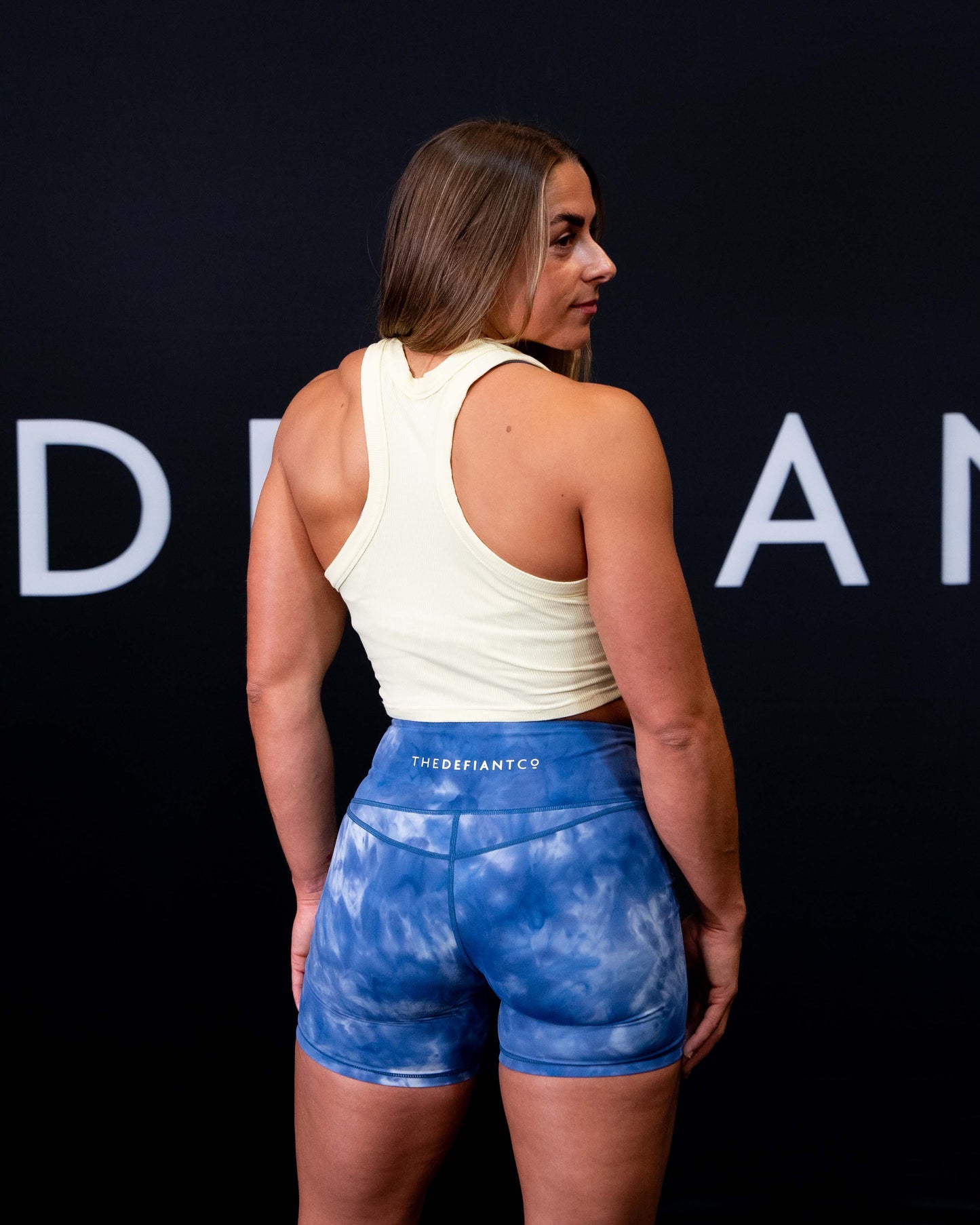 A girl against a black The Defiant Co backdrop wearing the DFNT. Ribbed Cropped Tank.  The tank is breathable with a stretchy fabric so is perfect for working out in.  It has a subtle DFNT. logo on a tag.  The colour is lemon.