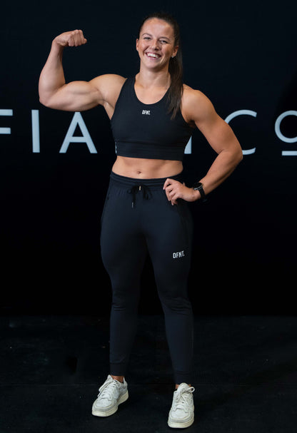 A girl against a black The Defiant Co backdrop wearing the DFNT. Black Jogging Bottoms and black sports bra.   It has a subtle DFNT. logo under the pocket. It has a drawstring waist and the pockets have zips. She is posing with a tensed arm.