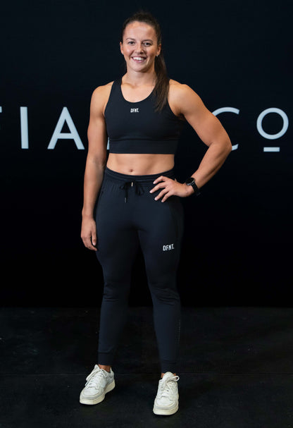 A girl against a black The Defiant Co backdrop wearing the DFNT. Black Jogging Bottoms and black sports bra.   It has a subtle DFNT. logo under the pocket. It has a drawstring waist and the pockets have zips. 