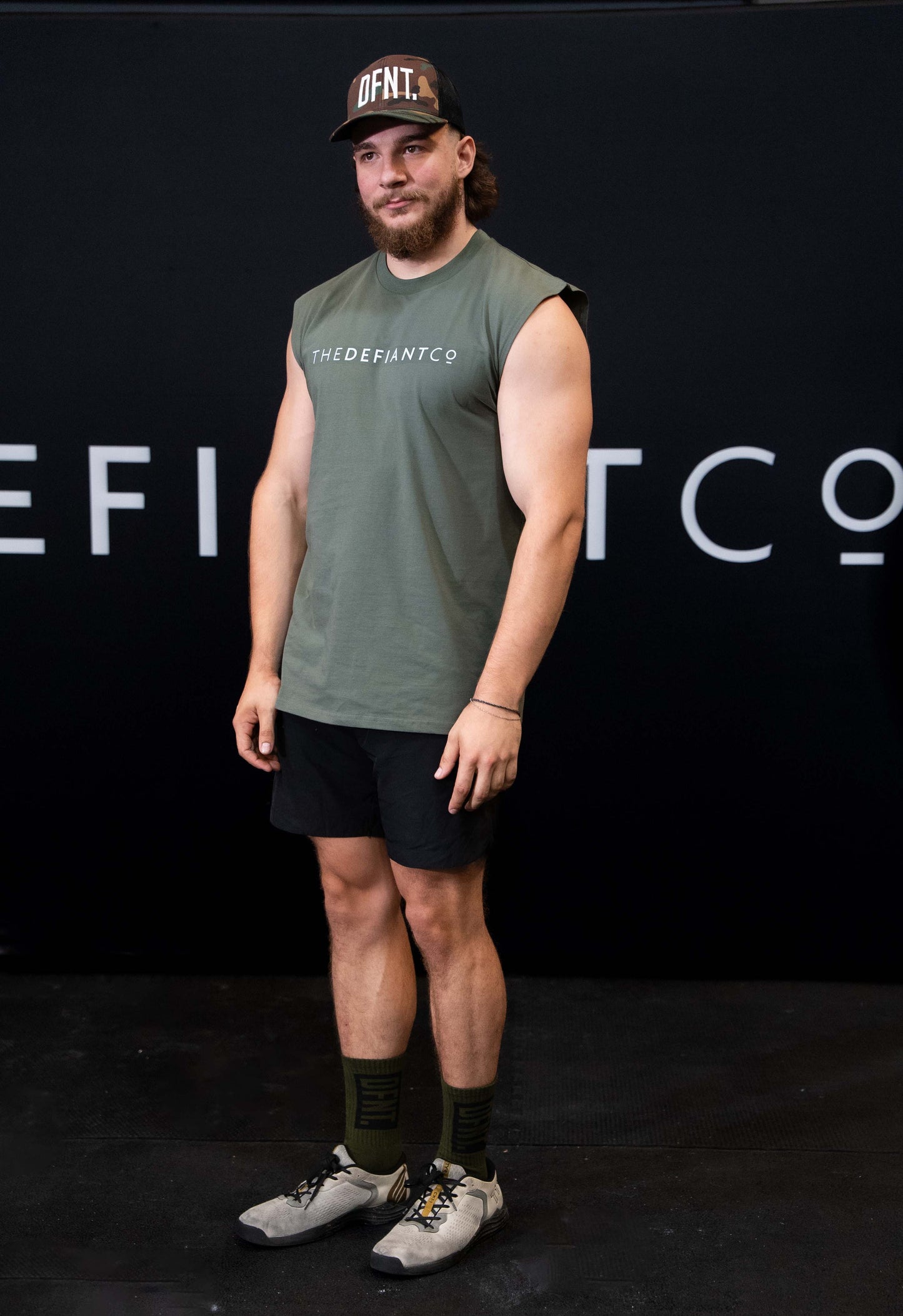 A man against a black The Defiant Co backdrop wearing the DFNT. Camo Trucker hat, Black TDCo performance shorts and egreen TDCo sleeveless tee.