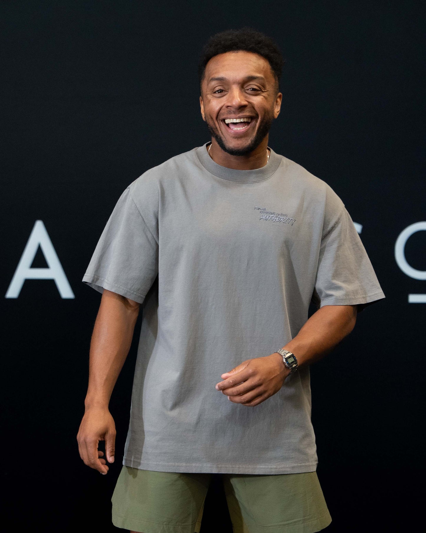 A guy against a black The Defiant Co backdrop wearing the faded grey proudly refusing to obey authority oversized tee and olive green performance shorts. 