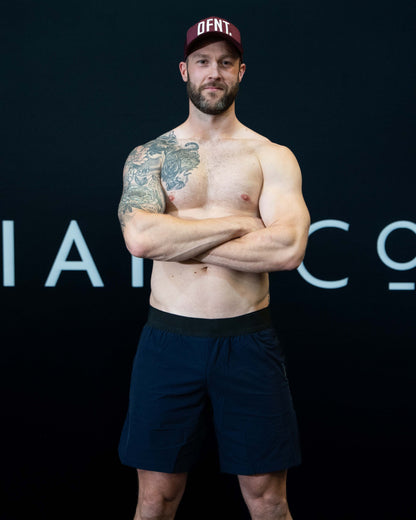 A man against a black The Defiant Co backdrop wearing the performance shorts with their draw string and elasticated black waist band.  The Defiant Co is subtly embossed down the right leg. The colour is navy blue.