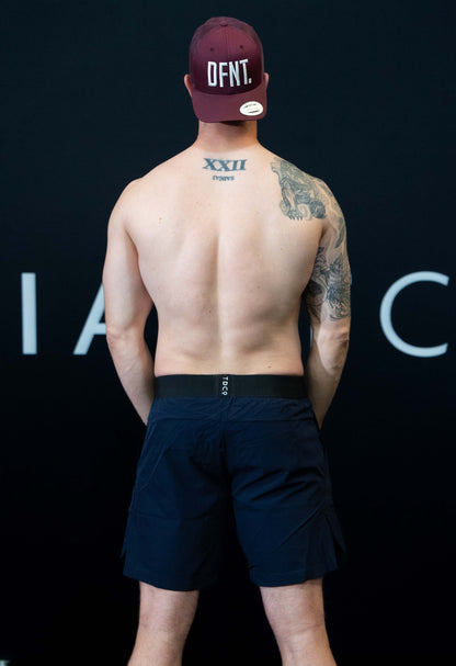 A man against a black The Defiant Co backdrop wearing the performance shorts with their draw string and elasticated black waist band.  The Defiant Co is subtly embossed down the right leg. The colour is navy blue.