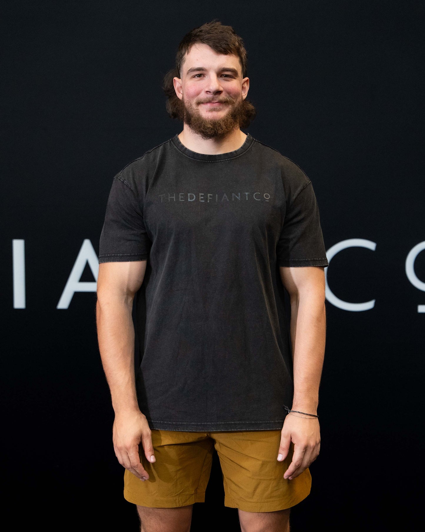 A man against a black The Defiant Co backdrop wearing the performance shorts with their draw string and elasticated black waist band.  The Defiant Co is subtly embossed down the right leg. The colour is gold.