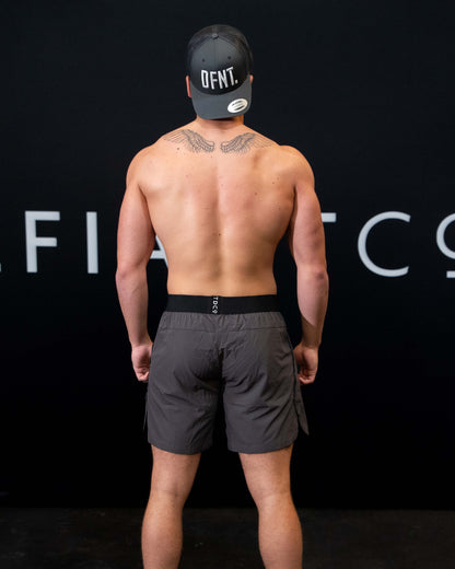 A man against a black The Defiant Co backdrop wearing the performance shorts with their draw string and elasticated black waist band.  The Defiant Co is subtly embossed down the right leg. The colour is charcoal grey.