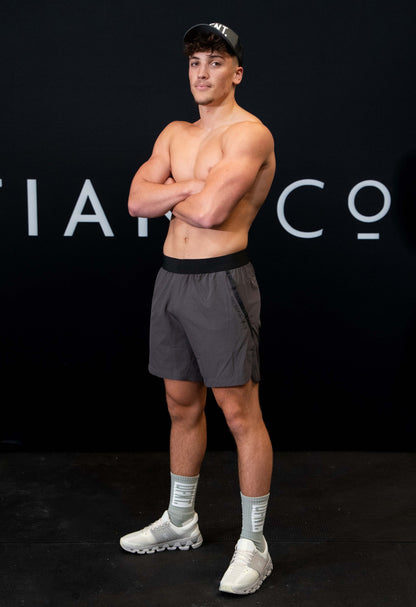 A man against a black The Defiant Co backdrop wearing the performance shorts with their draw string and elasticated black waist band.  The Defiant Co is subtly embossed down the right leg. The colour is charcoal grey.