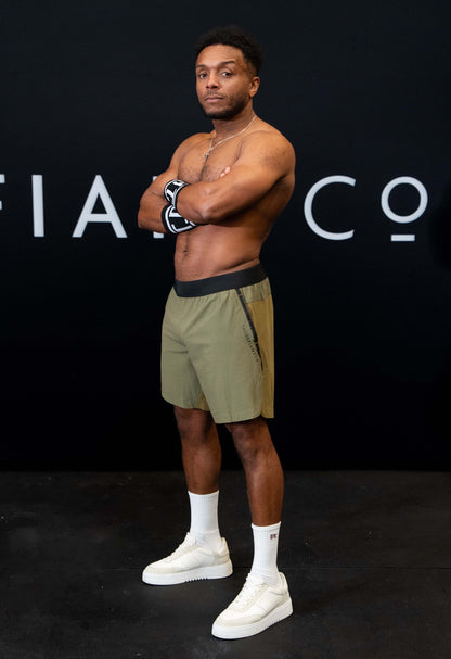 A man against a black The Defiant Co backdrop wearing the performance shorts with their draw string and elasticated black waist band.  The Defiant Co is subtly embossed down the right leg. The colour is olive green.