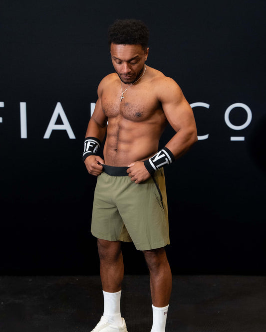 A man against a black The Defiant Co backdrop wearing the performance shorts with their draw string and elasticated black waist band.  The Defiant Co is subtly embossed down the right leg. The colour is olive green.