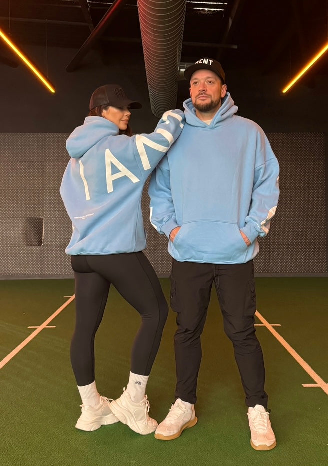 A guy and a girl stood posing in front of a church on a cloudy day, both dressed in the amazing Oversized DFNT. Eternity Hoodies. The Hoodies have a plain front with a big DEFIANT lettering across the arms and back, a subtle DFNT. tag on the left side and then embroidery stating Proudly Refusing To Obey Authority under the large lettering as well as The Defiant Co embroidered to the left sleeve cuff. The hoodies are light blue with white logos.