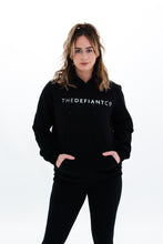 Load image into Gallery viewer, A girl wearing a standard fit, unisex The Defiant Co Hoodie.  The hoodie has draw strings and a pouch pocket on the front as per all standard designs.  The hoodie has the famous The Defiant Co logo across the front of the chest and is plain on the back.  The colour is black.